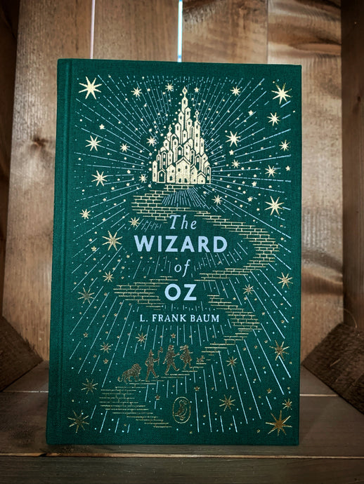 Image of the Puffin clothbound classic book with a front cover design featuring a forest green background and a gold foil printed image of the Emerald City at the end of the yellow brick road. The characters Dorothy, Scarecrow, Tin Man and the Cowardly Lion can be seen walking along the road all surrounded by gold stars and white rays of light.