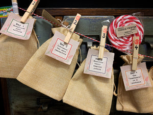 Image shows part of a Festive Countdown Calendar: four jute drawstring bags hanging from red and white striped twine, attached by pegs that are numbered from one to four and stamped with the Grimm and Co. insignia. Also pegged to the bags are small pieces of paper showing days one to four of the story writing challenge. Poking out from inside the bags are a Rainbow Word Wand, Leprechaun wishes, a piece of Atlantean Sea Glass, and a Tongue Twister lollipop.