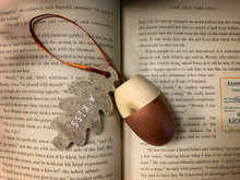 Load image into Gallery viewer, Image of A Kiss, a wooden acorn-shaped hanging ornament with a natural wood finish on the top half, and rose gold painted bottom half. Attached to the hanging loop is a grey felt oak leaf with the words A Kiss printed in white. Decoration is shown displayed on the pages of Peter Pan.