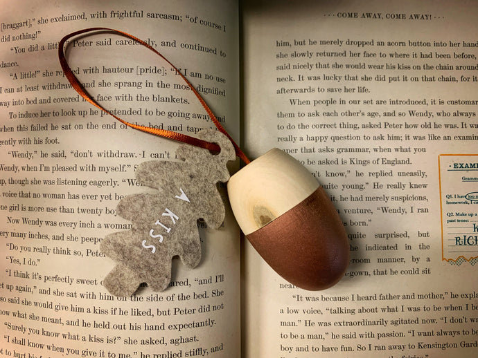 Image of A Kiss, a wooden acorn-shaped hanging ornament with a natural wood finish on the top half, and rose gold painted bottom half. Attached to the hanging loop is a grey felt oak leaf with the words A Kiss printed in white. Decoration is shown displayed on the pages of Peter Pan.