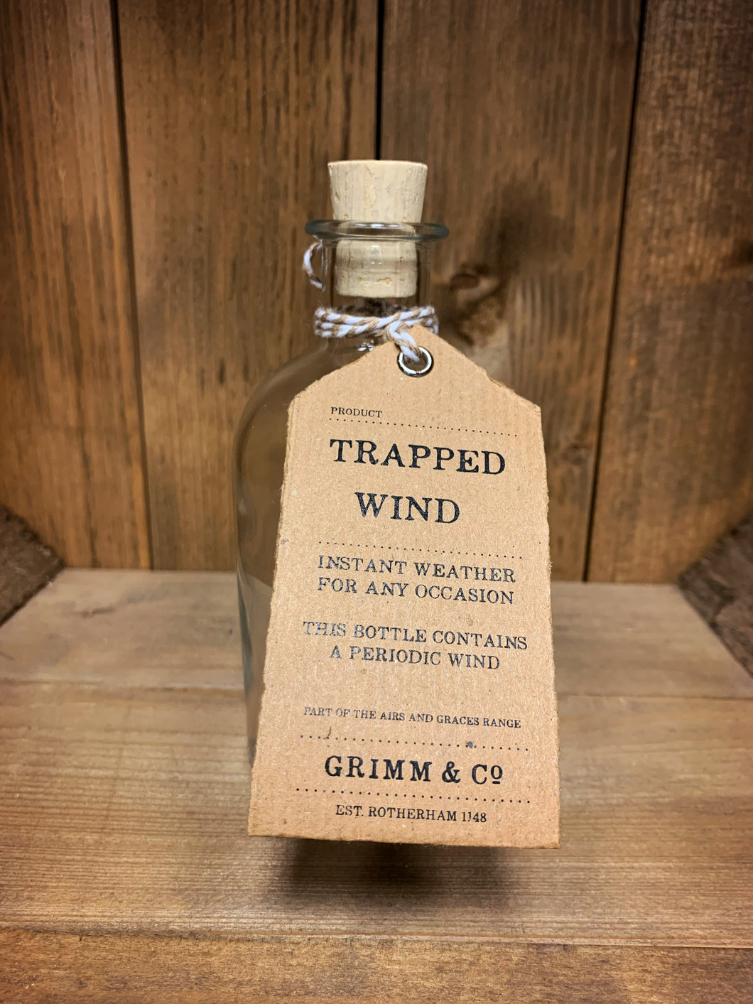 Image of the Trapped Wind bottle from the Airs and Graces range: An empty glass potion bottle with cork. The bottle has a kraft tag around  the neck, reading: Trapped Wind. Instant weather for any occasion. This bottle contains a periodic wind. 