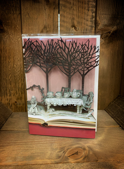 Image of an A5 laser cut greetings card with a pink background (inside the card). The design is cut to show an open book with the pages of the story crafted into the Mad hatter's tea Party scene with trees overhead and Alice stood on the page.