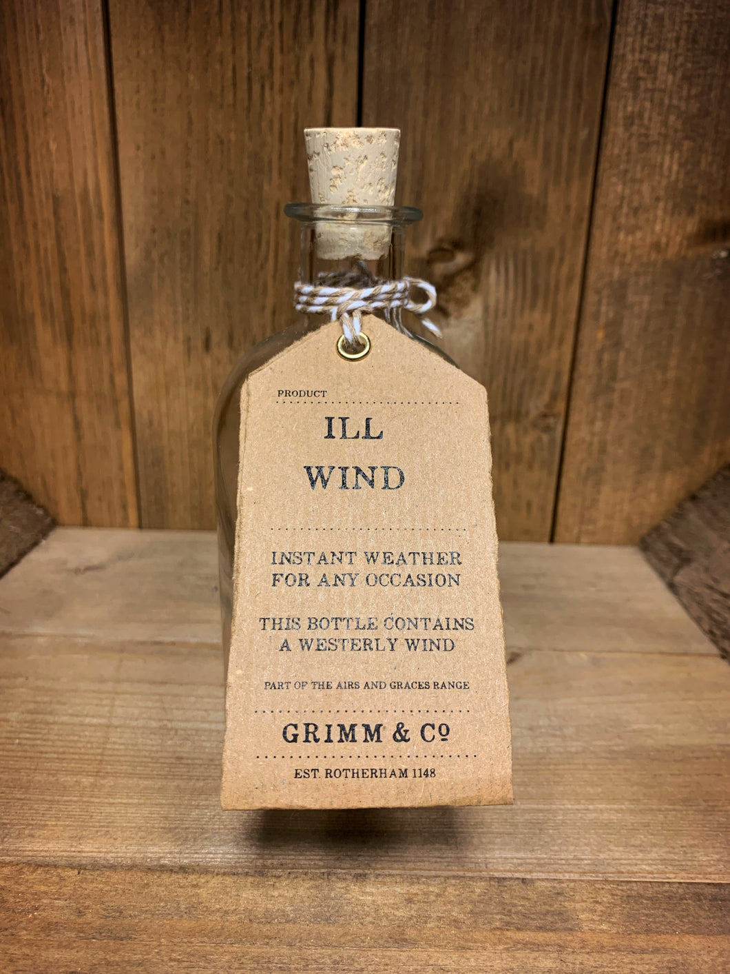 Image of the Ill Wind bottle from the Airs and Graces range: an empty glass potion bottle with cork. The bottle has a kraft tag around the neck, reading: Ill Wind. Instant weather for any occasion. This bottle contains a westerly wind. 