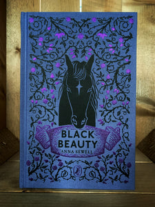 Image of the front cover of the Puffin clothbound classic Black Beauty with a purple background and a close-up print of a black horse and rose thorns with purple foiled roses and buds in a mirrored pattern.