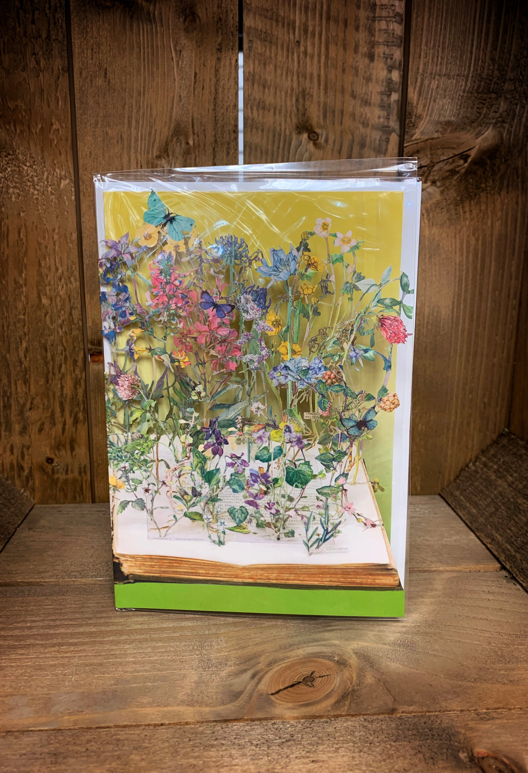 Image of an A5 laser cut greetings card with a yellow background (inside the card) and a cut out scene popping up from an open book showing wildflowers in various colours and a blue butterfly in the top corner.