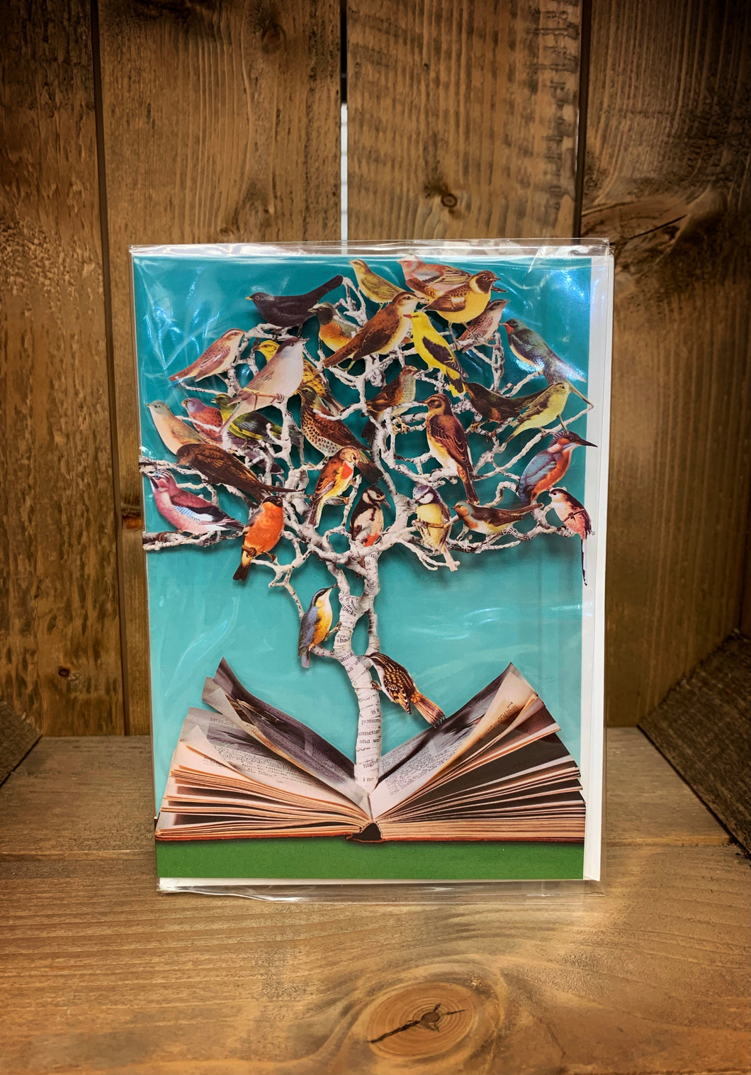 Image of an A5 laser cut greetings card with a turquoise background (inside the card). The card has a laser cut tree coming up from the centre pages of an open book and nestled in the tree are all different kinds of birds in the branches.
