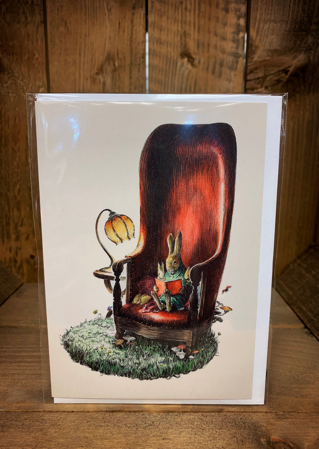 Image showing a cream coloured A6 greetings card with a design showing a family of rabbits sat curled up in a red armchair whilst the largest rabbit reads from a red book. There is grass with toadstools growing underfoot and a natural shaped lamp to read by conjuring up ideas of cosy bedtime stories. Background of the cad is cream coloured.
