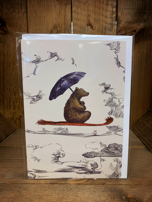 Image of a cream A6 greetings card showing a brown bear sat cross-legged on a red flying carpet soaring across the cloudy sky whilst holding a blue umbrella. 