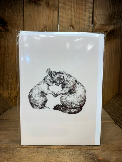 Image shows a cream A6 greetings card with a design showing a black and white sketch of a rabbit and a cat sat facing each other with heads bowed together and paws touching as though in a hug. 