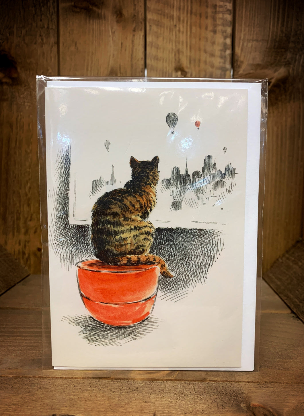 Image of a cream A6 greetings card showing a design with a tabby cat sat on a red stool facing a window overlooking a city scene in the distance and three hot air balloons rising in the sky