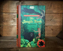 Load image into Gallery viewer, Image showing the book-shaped Jigsaw Library The Jungle Book with a front cover design featuring Mowgli swinging on a vine with Baloo the bear and Bagheera and Kaa in the trees above. The shadow in Shere-Khan can be seen in the background.