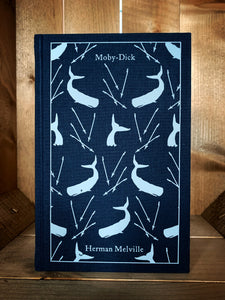 Image of the front cover of the Penguin clothbound classic Moby Dick featuring a navy blue background and a repeat pattern of pale grey whales whale tails with harpoons interspersed among them.