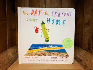 Image shows the front cover of the board book The Day The Crayons Came Home with Pea Green crayon standing on a postcard with a knapsack over their shoulder. 