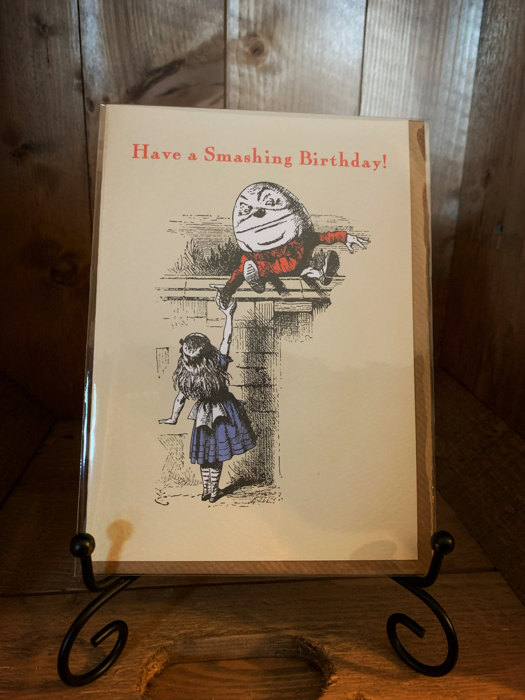 Image shows the greetings cards Alice Humpty Dumpty Birthday with a taupe background and a black and white illustration of Alice with Humpty Dumpty wearing blue and red outfits and the words 'Have a Smashing Birthday!' across the top