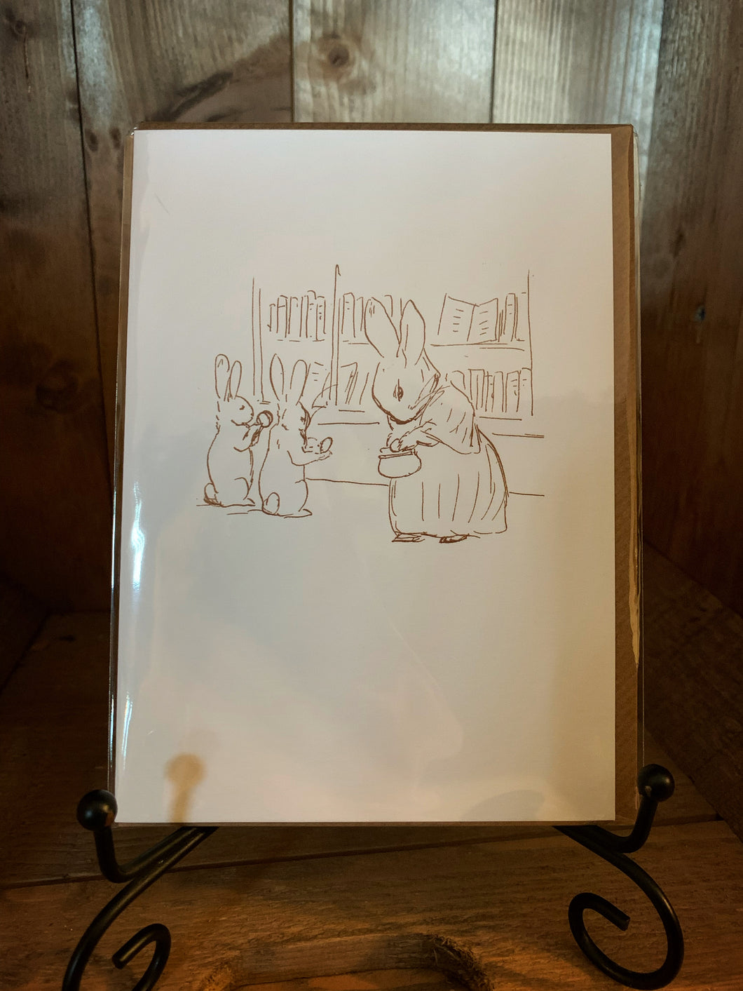 Image of the greetings card Beatrix Potter Rabbits Shopping with a cream background and a brown linear illustration of a mother rabbit and two little rabbits shopping for books