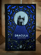 Load image into Gallery viewer, Image of the front cover of the Puffin clothbound classic of Dracula. The cover features a black background with the silhouette of a castle against a white moon atop a rocky mountain printed in purple foil and surrounded by purple foil bats and white stars.