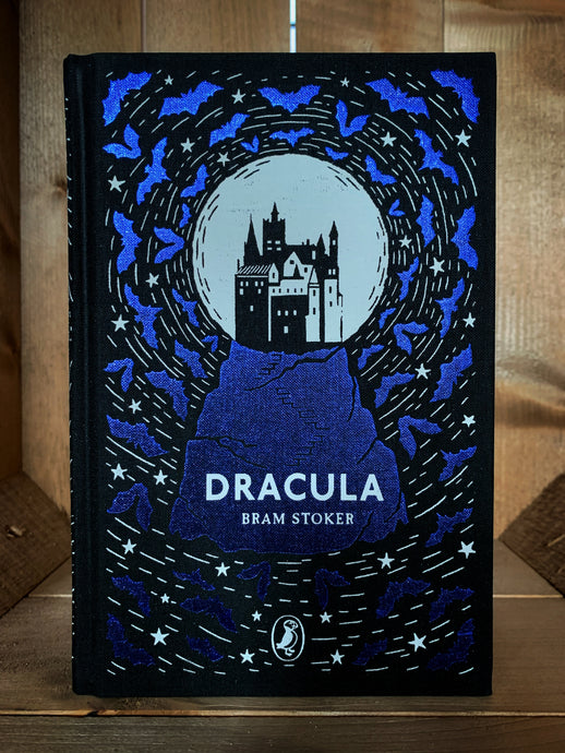 Image of the front cover of the Puffin clothbound classic of Dracula. The cover features a black background with the silhouette of a castle against a white moon atop a rocky mountain printed in purple foil and surrounded by purple foil bats and white stars.
