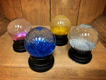 Load image into Gallery viewer, Image of the four types of glitter globe (snow globes full of coloured glitter, with a black base).  The image shows the globes after being shaken. From left to right: pink, blue, gold, and silver. 