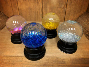 Image of the four types of glitter globe (snow globes full of coloured glitter, with a black base).  The image shows the globes after being shaken. From left to right: pink, blue, gold, and silver. 