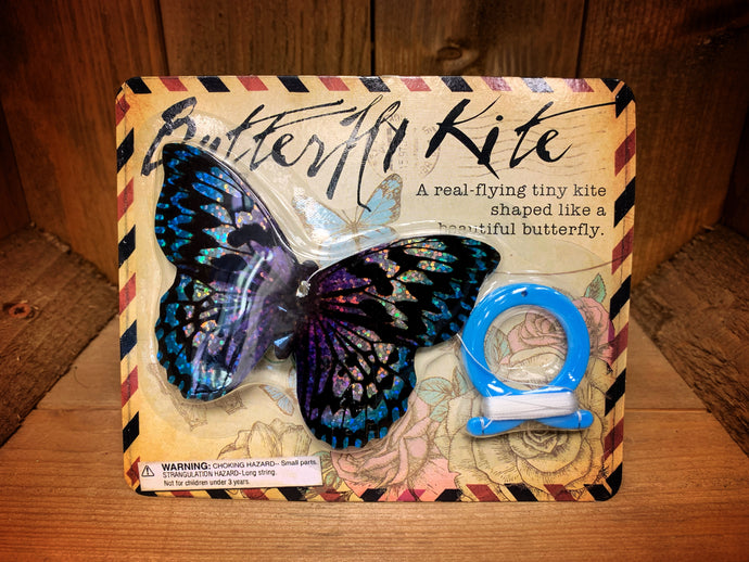 Image shows a mini butterfly kite in it's packaging. The kite in this image has a blue, purple, and black iridescent butterfly wing pattern.
