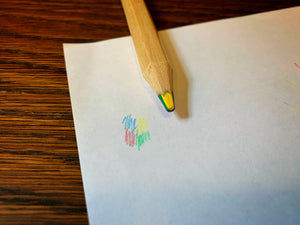 Image shows a close up of the lead in a Rainbow Word Wand. The pencil is resting on a piece of white paper, next to a small scribbled example of the four included colours that make up the lead: blue, yellow, red, and green.