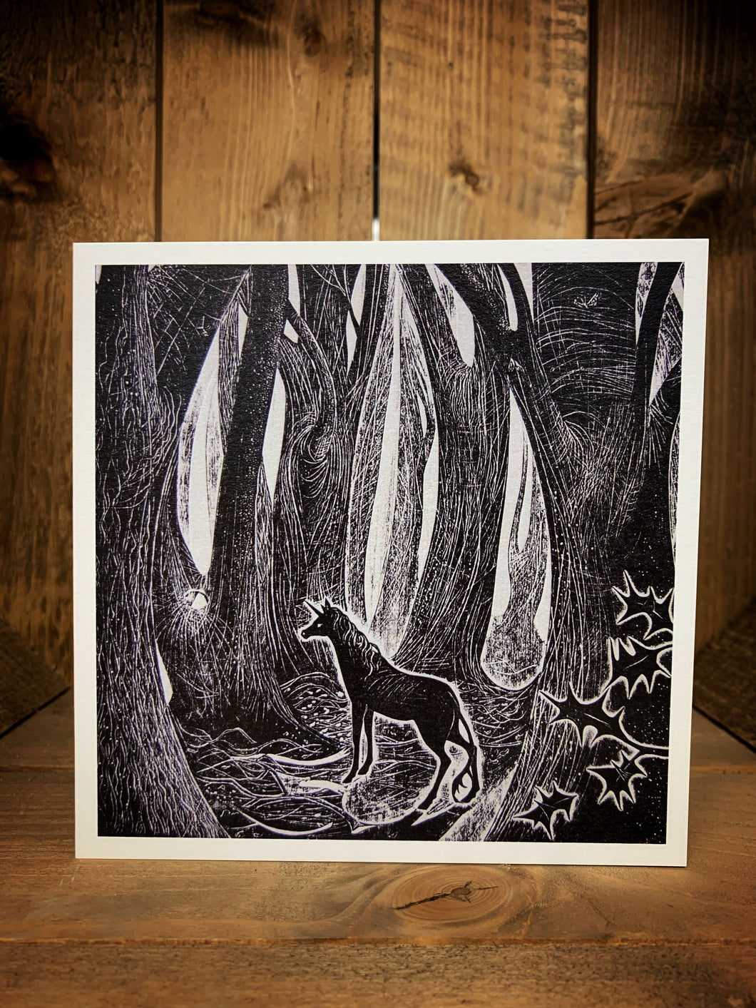 Image of a square greetings card featuring a black and white image block printed showing a forest scene with a unicorn in a clearing with a white border and holly leaves in the foreground.