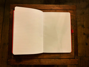 Image is of a Red Note Keeper laid out open, showing the lined pages. The likes on the page are slightly slimmer than a standard lined notebook. The elasticated pen holder can be clearly seen attached on the right side in the middle.