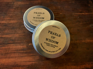 Image shows two tins of Pearls of Wisdom. Two small, round, aluminium screw-top tins with a kraft sticker label, one flat down on a surface, with the other leaning on it. 
