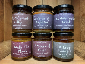 Image of all six types of chutney and preserves stacked in rows of three. On the bottom row: Walk the Plank, A Shred of Hope, A Love Triangle. On the top row: The Slightest Folly, A Sense of Deja Vu,  An Anticipated Event. All are in small cylindrical jars, with a dark, blackboard style label, with cursive white text, and black lids.