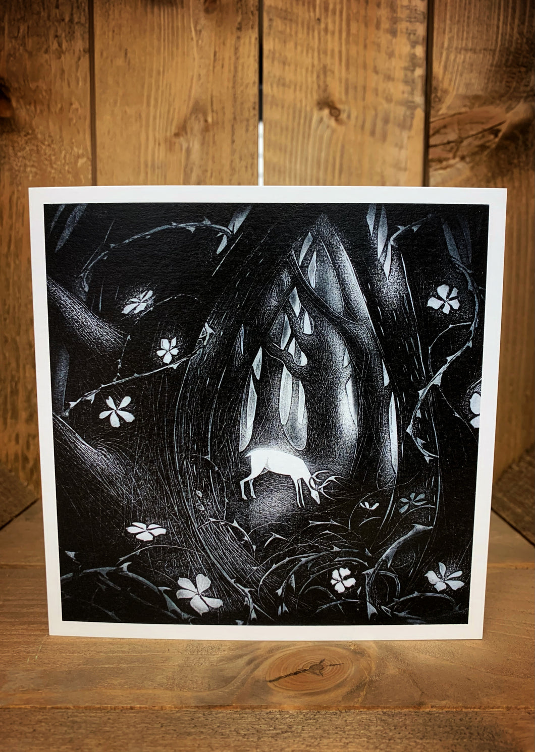Black and white square greetings card showing a forest with a white stag in the clearing and white roses and thorns i the foreground.