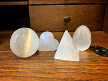 Load image into Gallery viewer, Image shows the four shape variants of Blue Moon Crystal (selenite crystal). From left to right: sphere, heart, pyramid, and egg.  They are resting on a wooden surface.