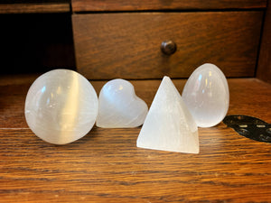 Image shows the four shape variants of Blue Moon Crystal (selenite crystal). From left to right: sphere, heart, pyramid, and egg.  They are resting on a wooden surface.