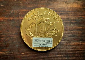 Image shows a single Dragon's Hoard chocolate coin - a giant chocolate coin with a gold 'half-penny' embossed foil coating and a small kraft Grimm & Co. product sticker on the lower half. 