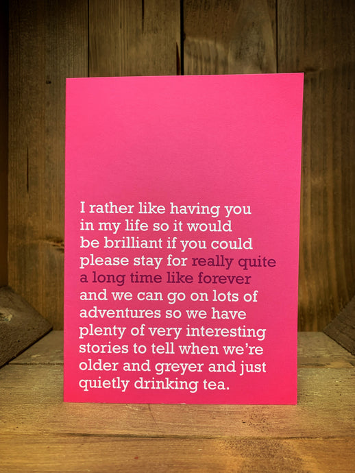 Image of the pink coloured greetings card with a typed message on the front that reads 'I rather like having you in my life so it would be brilliant if you could please stay for really quite a long time like forever and we can go on lots of adventures so we have plenty of interesting stories to tell when we're older and greyer and just quietly drinking tea.' The test is in white and dark pink with a blank inside.