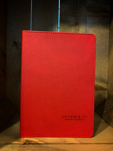 Image shows the front of a Red Note Keeper. An A5 notebook, with a red PVC cover. the cover has ‘Grimm & Co’ and ‘Apothecary to the Magical’ laser cut into the lower right hand corner.