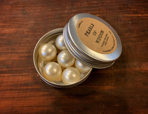 Image shows an open tin of Pearls of Wisdom, with the lid leaning against the tin. There are 7 cream-coloured bath pearls in the tin. 