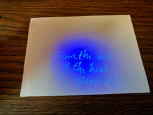 Load image into Gallery viewer, Image shows a white piece of paper being lit up with the blue UV light from the Invisible Ink Pen. Where the UV light hits, there is writing that reads: &#39;From the desk of the Head Shop Elf&#39;.