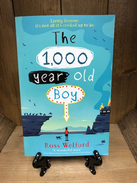 Image of the front cover of paperback book The 1,000 Year Old Boy featuring an illustration of a boy looking out across a landscape of blue with cliffs and a house in flames atop a cliff.