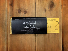 Load image into Gallery viewer, Image of A Blissful Interlude chocolate bar in Milk with a black label wrapped around gold foil. Bar is shown with printed golden ticket poking out the side of the wrapper.