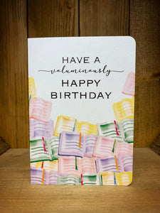 Image shows the front of the greetings card with a white background and illustrations of open books in green, purple, yellow and pink with the message 'Have a voluminously happy birthday'.